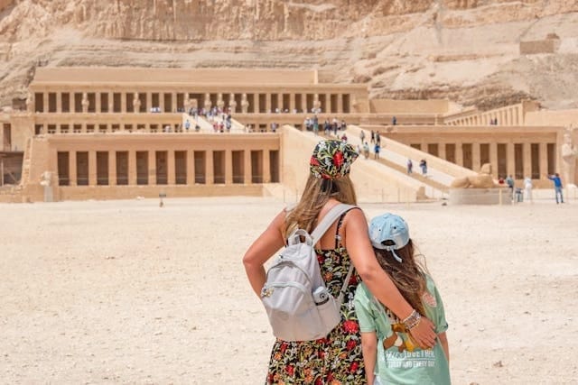 Eine Mutter und ihre Tochter in Sommerkleidung blicken auf den monumentalen Totentempel der Hatschepsut in Luxor während eines Ausflugs in den Sommermonaten.
